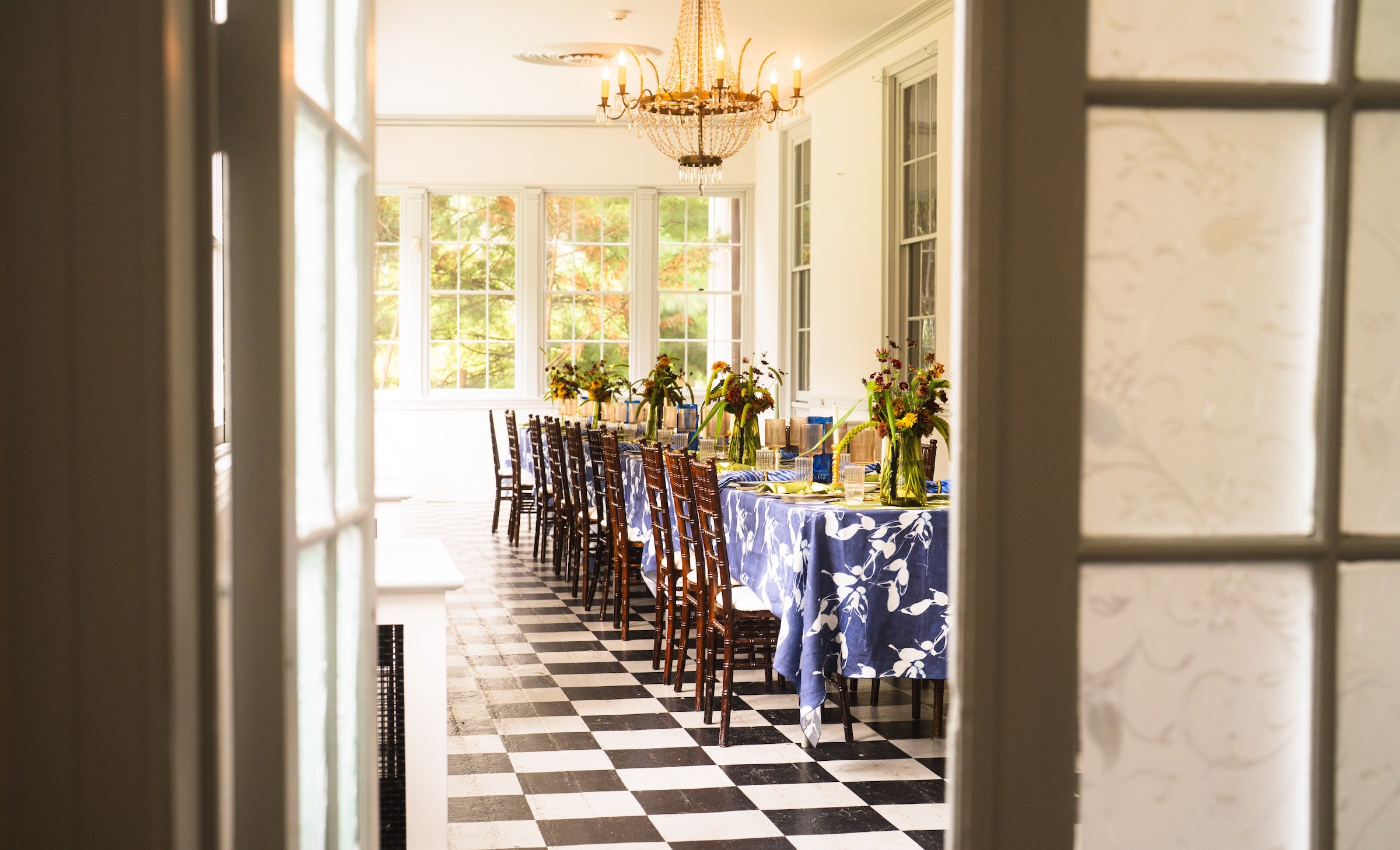 Long dining table set with chairs in a room with large windows and checkered floor.